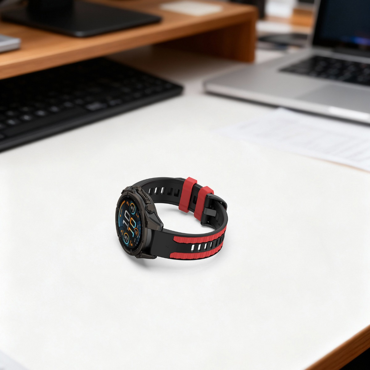 Eine schwarze Smartwatch mit rot-schwarzem Band liegt auf einem weißen Schreibtisch neben einem Laptop.