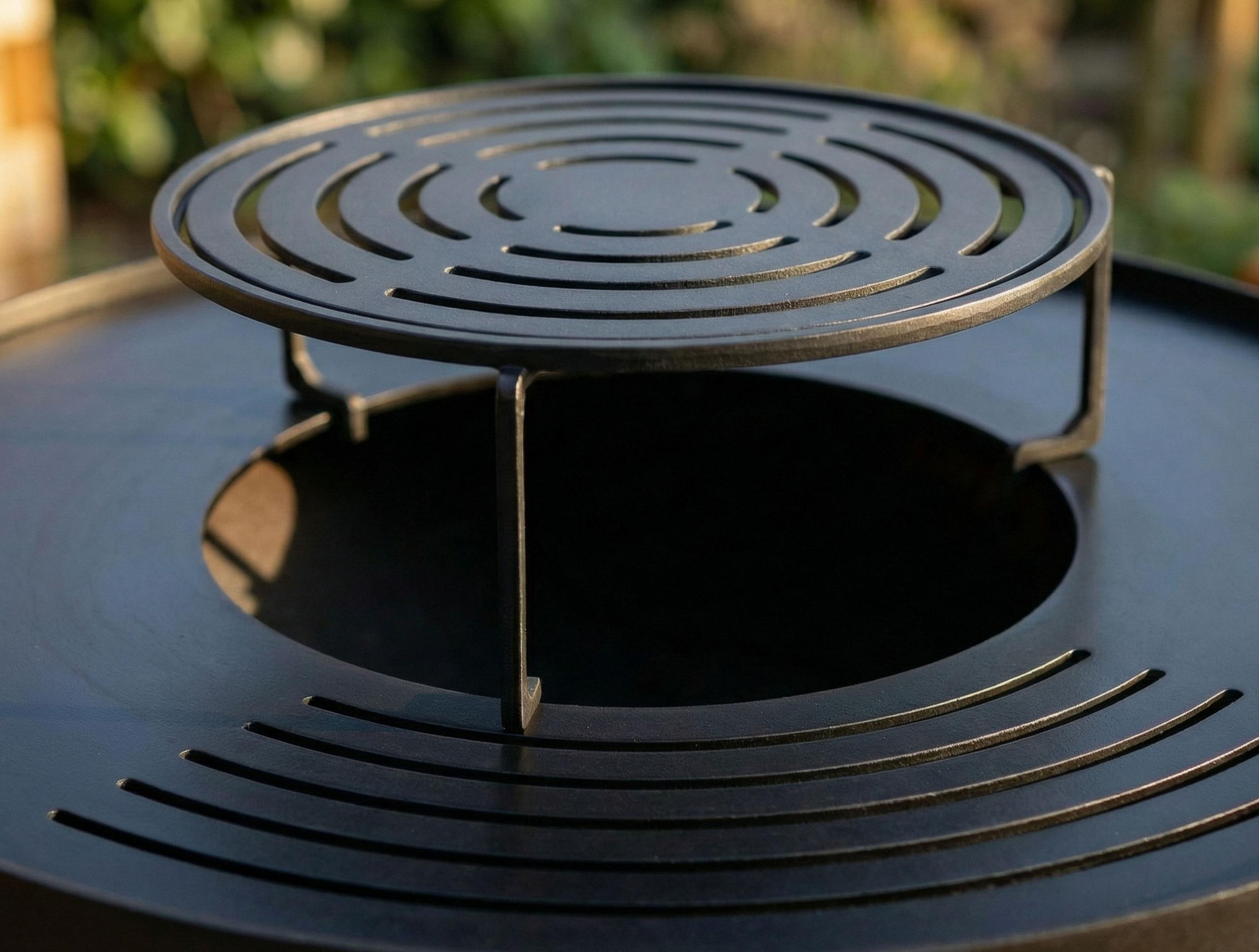 Ein runder, schwarzer Grillrost mit konzentrischen Schlitzen steht auf einem Grill.