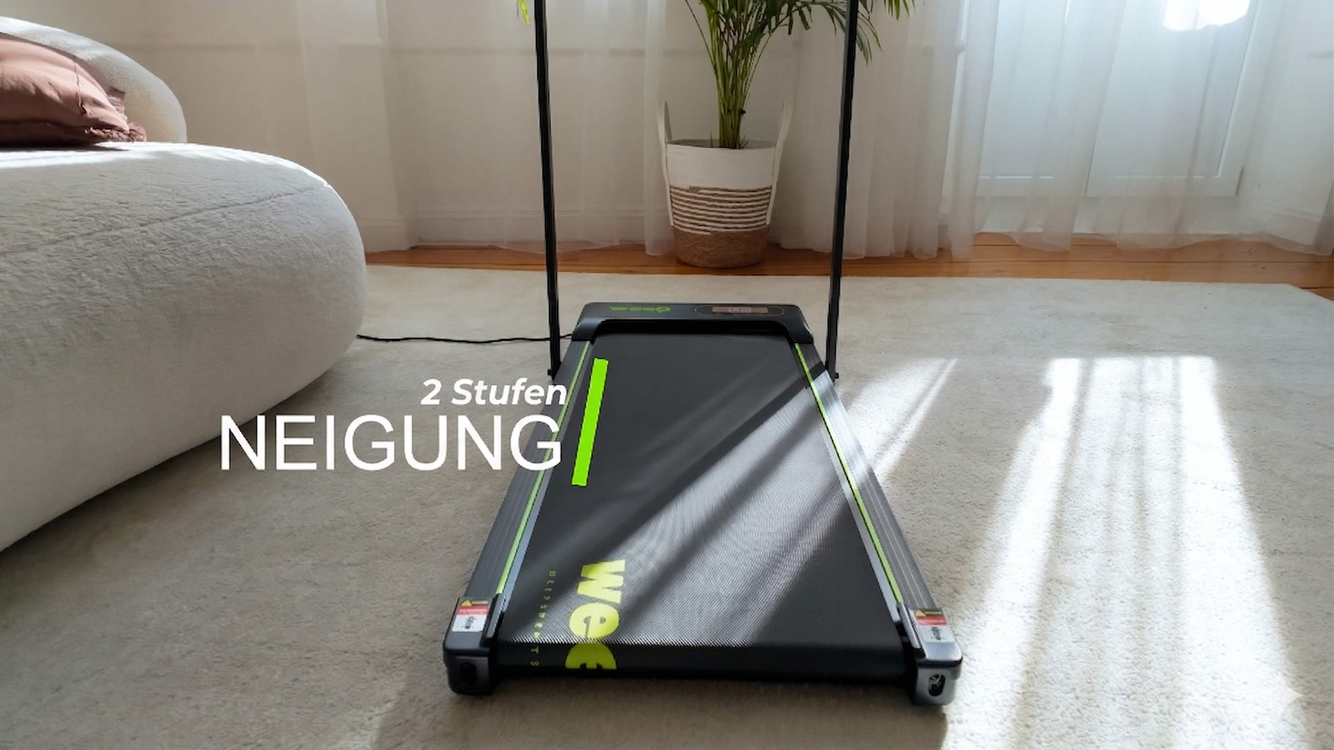 Bieżnia z zielonymi akcentami stoi na jasnym dywanie. Po lewej stronie kanapa.