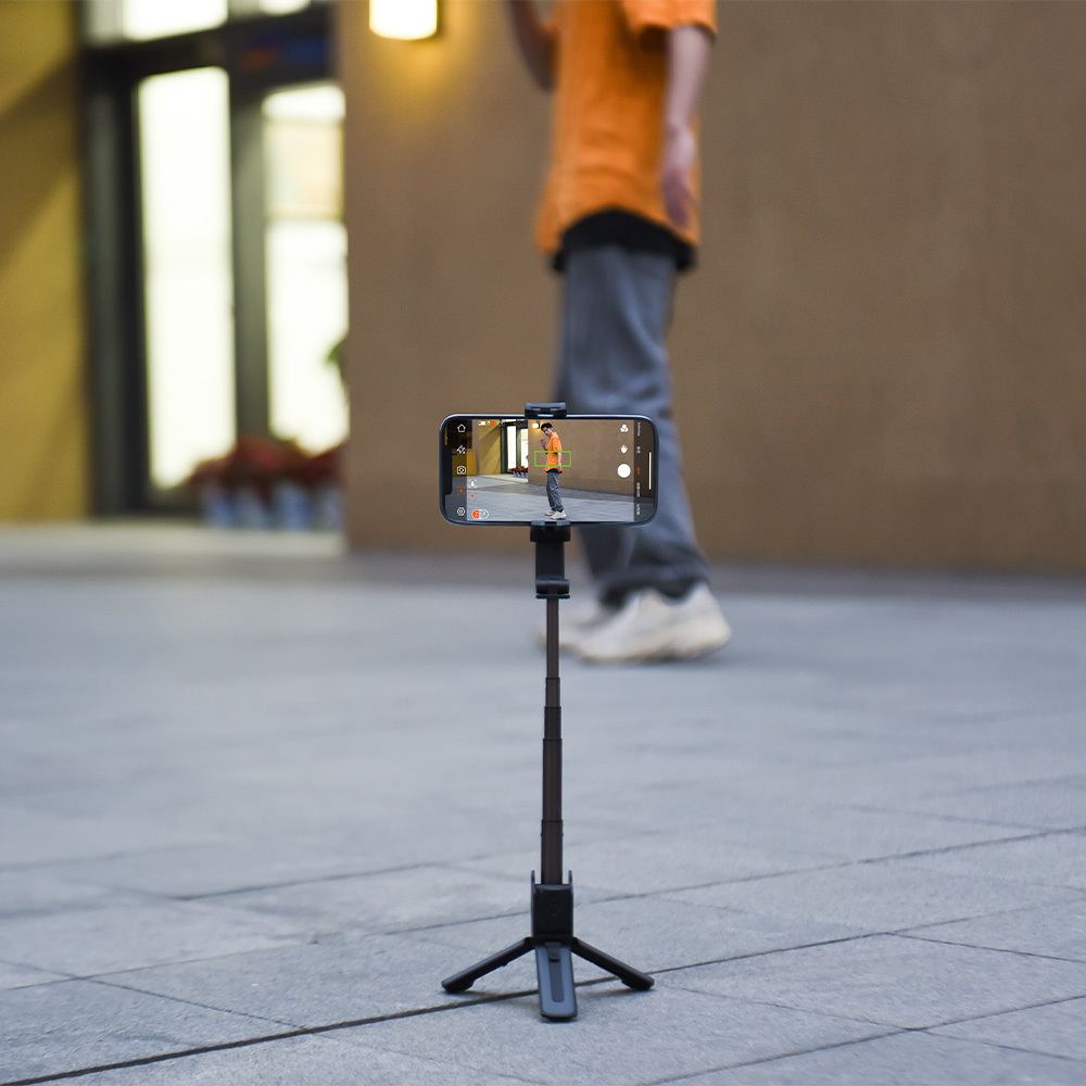 Smartfon na statywie z kijem do selfie filmuje osobę w pomarańczowej koszuli.