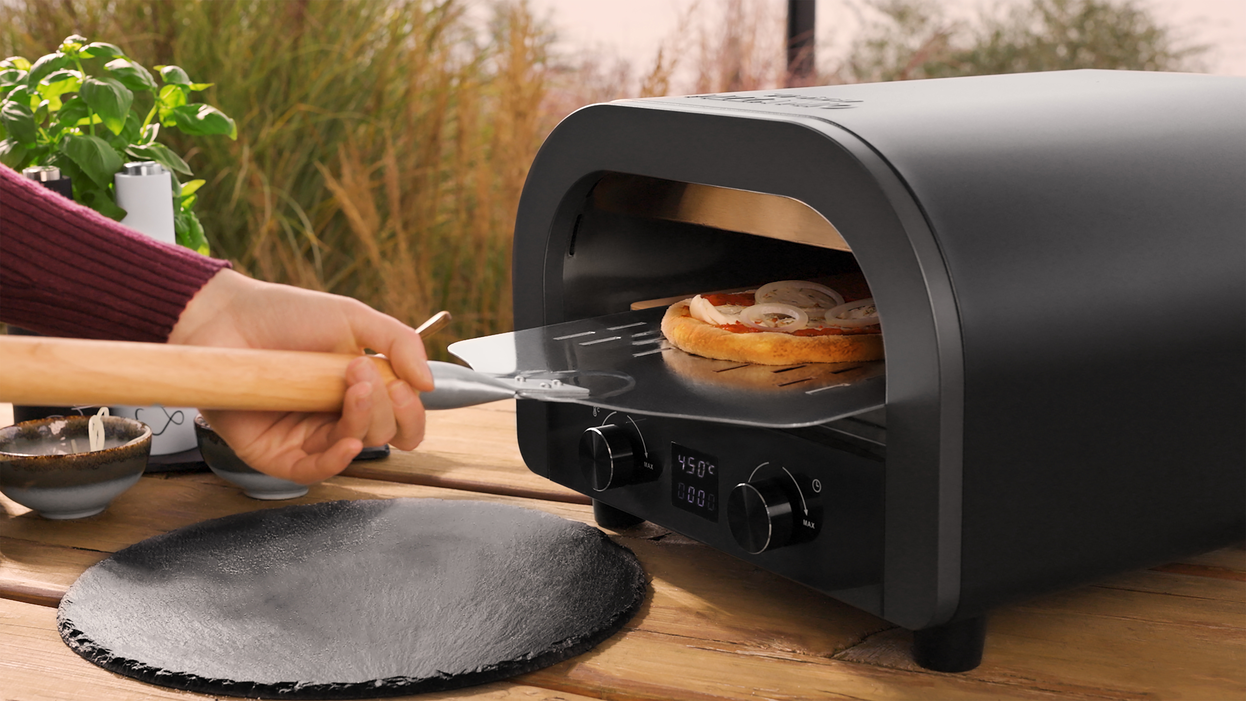 Eine Hand schiebt eine Pizza mit Zwiebeln in einen schwarzen Ofen auf einem Holztisch.