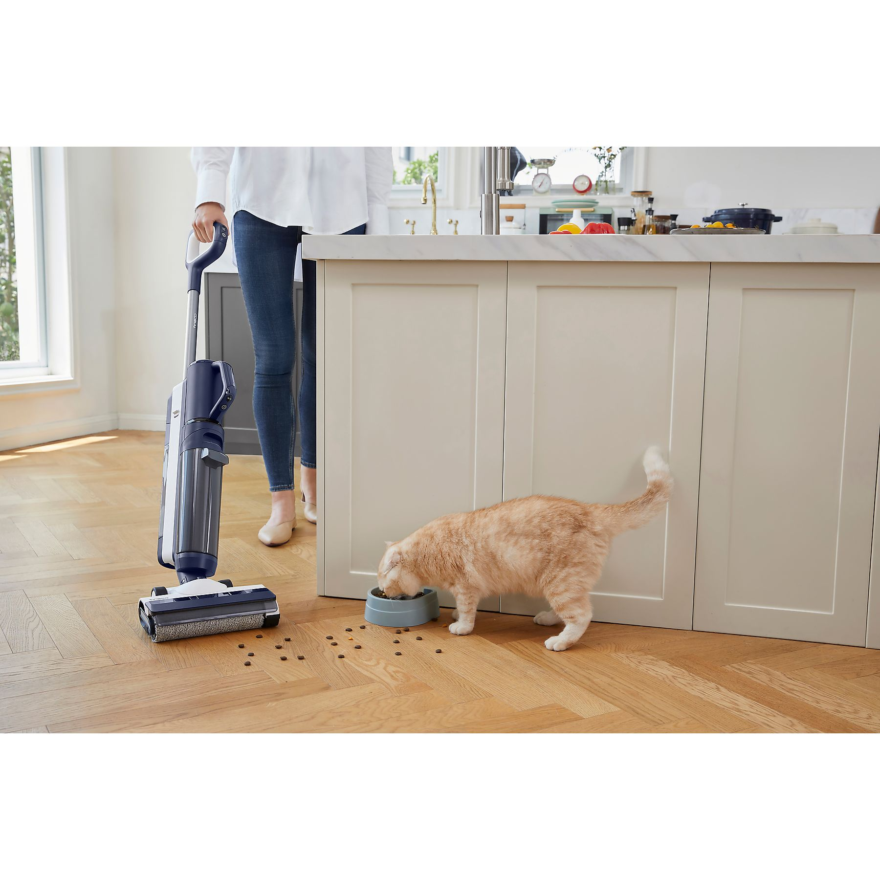 Eine Person benutzt einen Staubsauger, während eine Katze aus einem Napf auf Holzboden frisst.