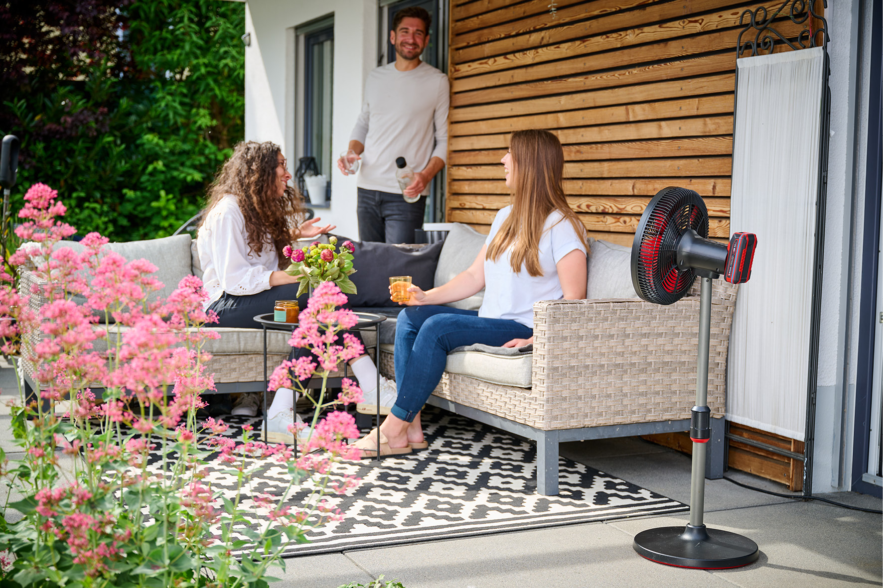 Tre persone su un patio con fiori e un ventilatore.