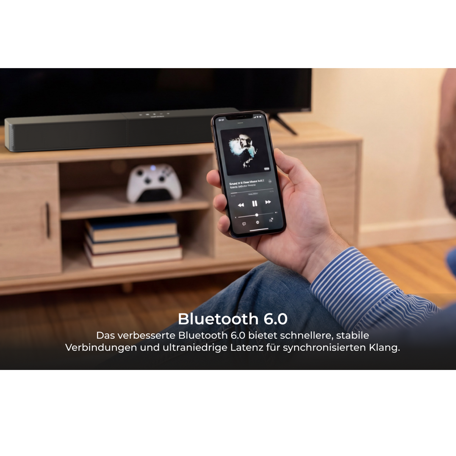 Hombre sostiene smartphone con reproductor de música frente a barra de sonido, mando y libros.