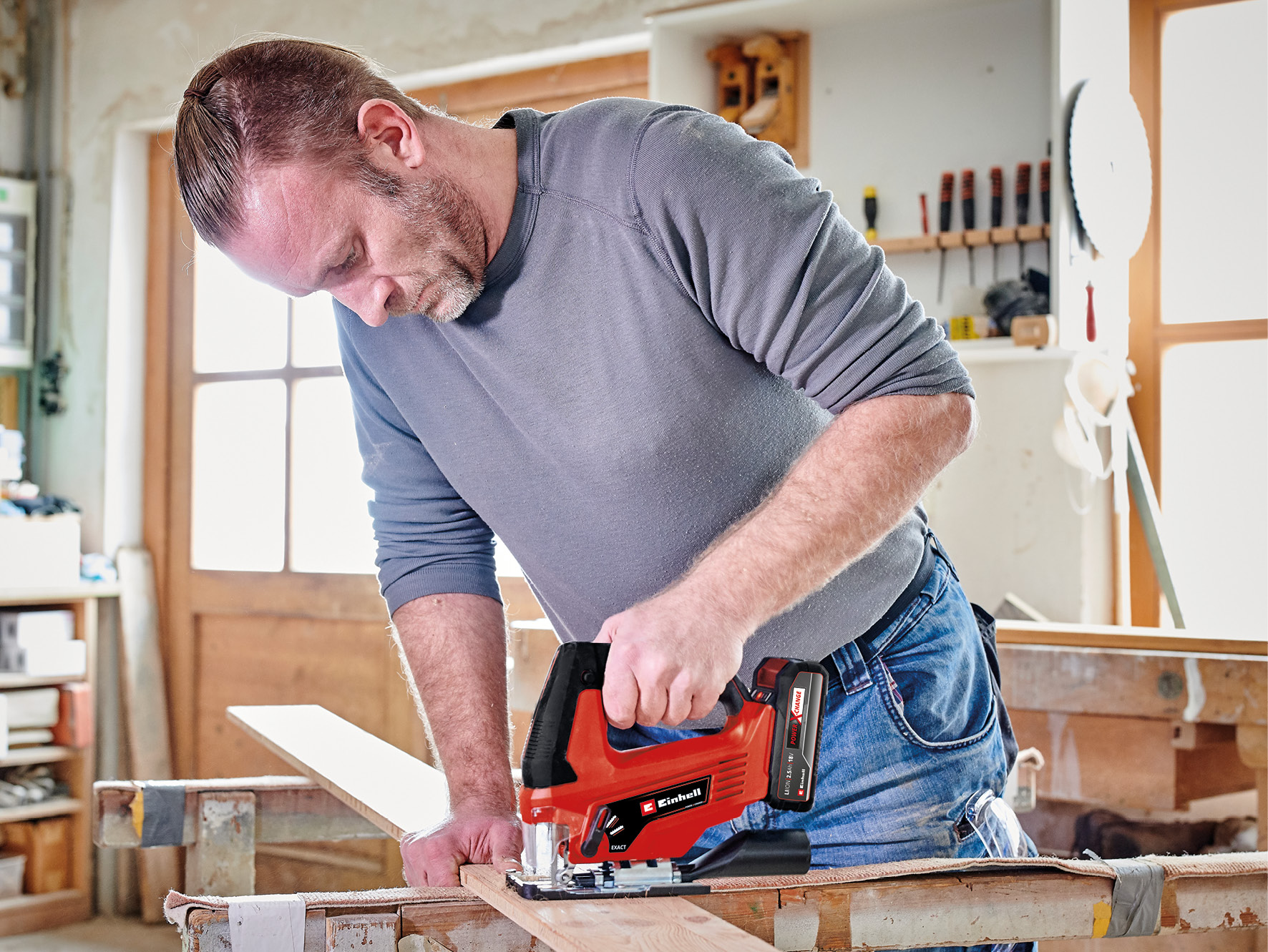 Mężczyzna z kucykiem i brodą tnie drewno wyrzynarką w warsztacie.