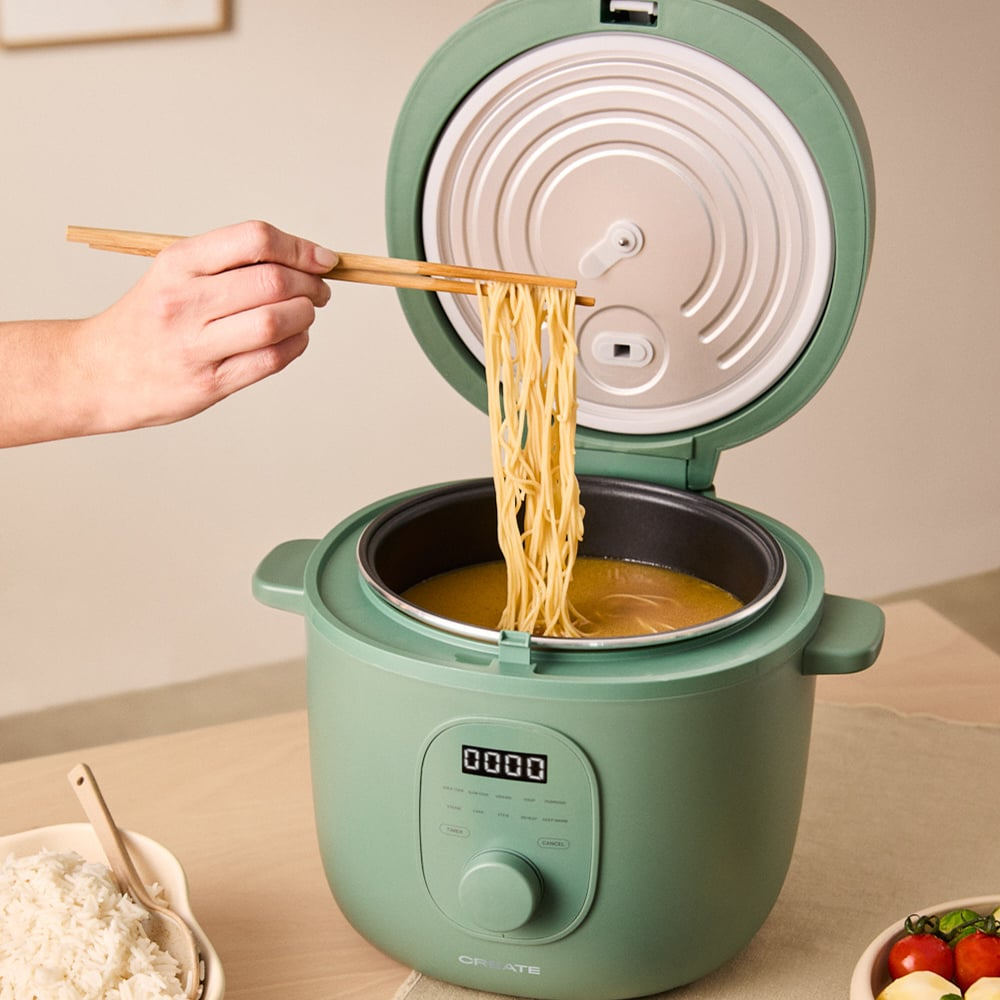 Una mano levanta fideos con palillos de una olla verde con caldo.