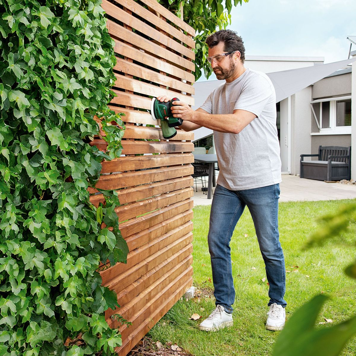 Mann schleift Holzlattenzaun mit Schleifmaschine im Garten. Ranken wachsen links.