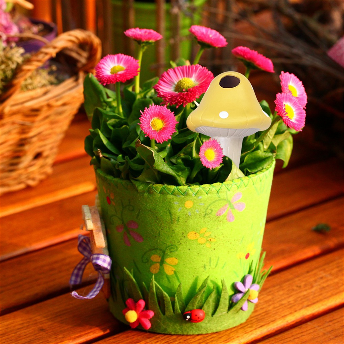 Ein grüner Topf mit rosa Gänseblümchen und einer gelben Pilzdekoration steht auf einem Holztisch.