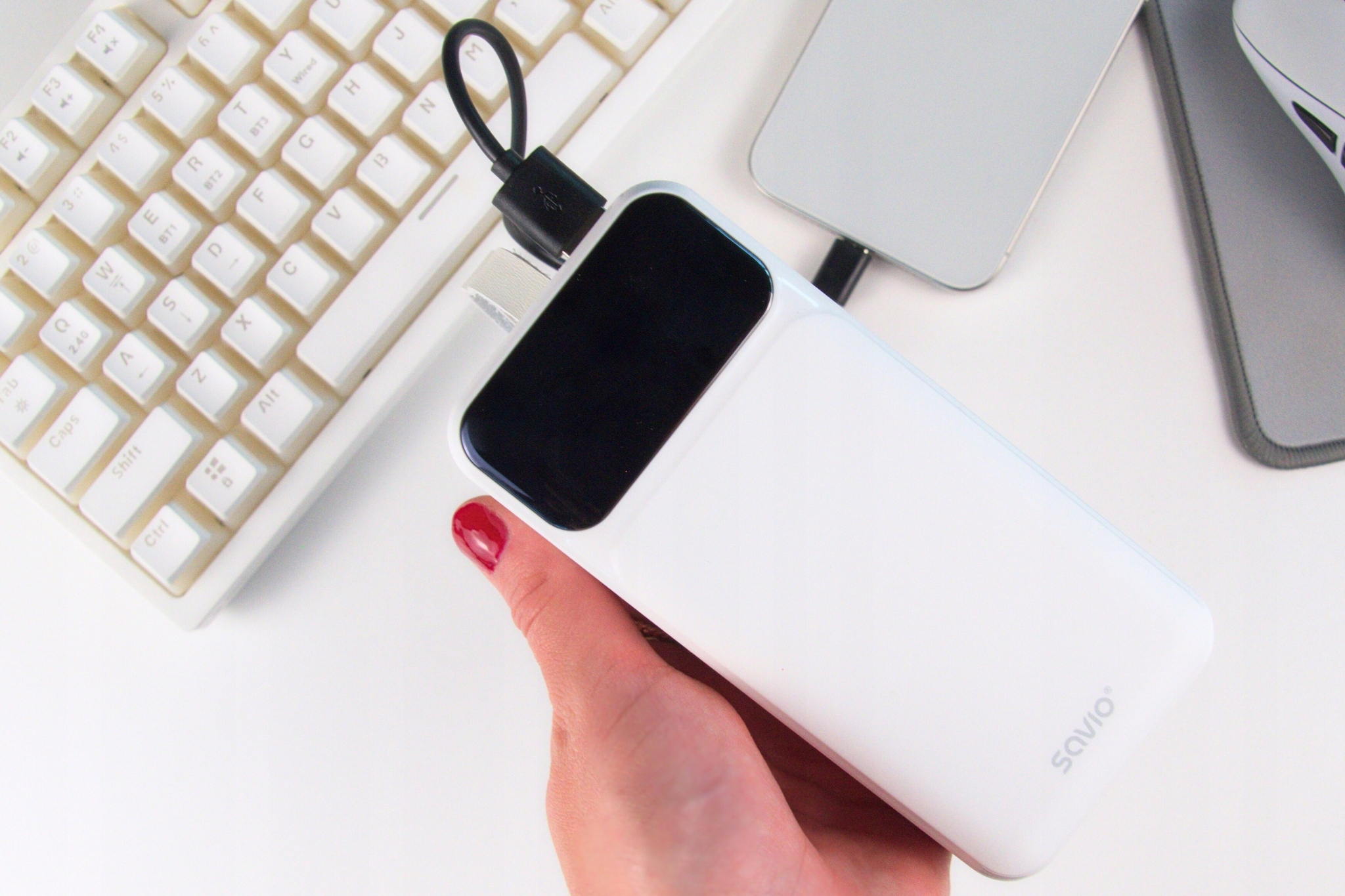 Dłoń trzyma biały power bank z czarnym wyświetlaczem, obok klawiatury i laptopa.