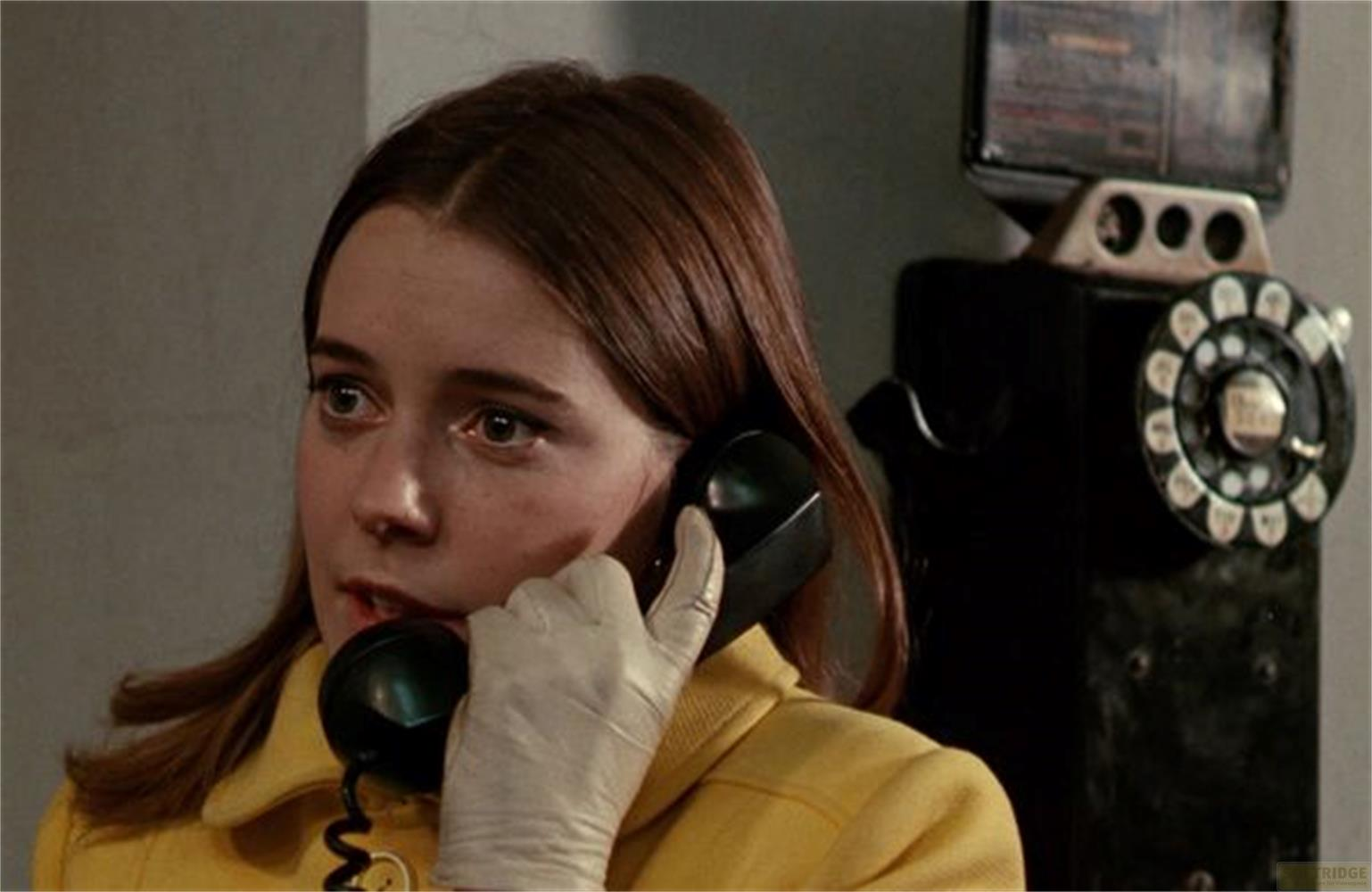 Una donna con i capelli castani indossa un cappotto giallo e guanti bianchi mentre parla al telefono.