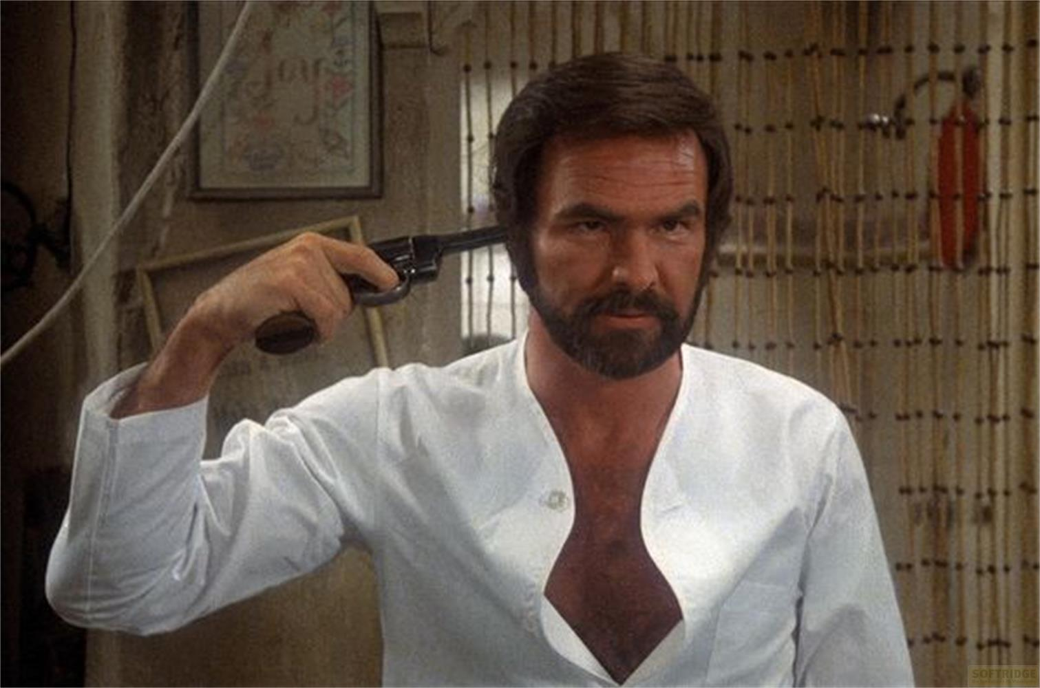 Un uomo con capelli scuri e barba tiene una pistola alla tempia.