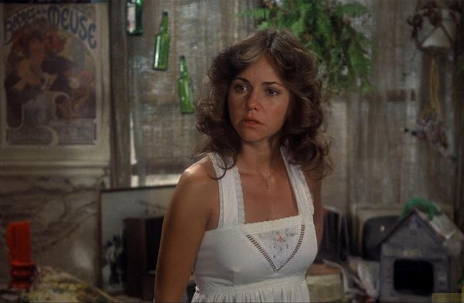 Una donna con capelli castani indossa un vestito bianco. Sullo sfondo ci sono bottiglie e un poster.