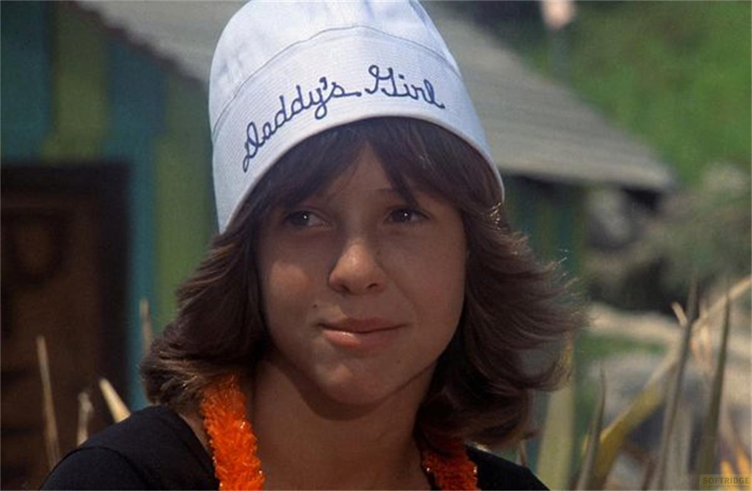 Una bambina dai capelli castani indossa un cappello bianco con scritto 'Daddy's Girl' e una collana arancione.