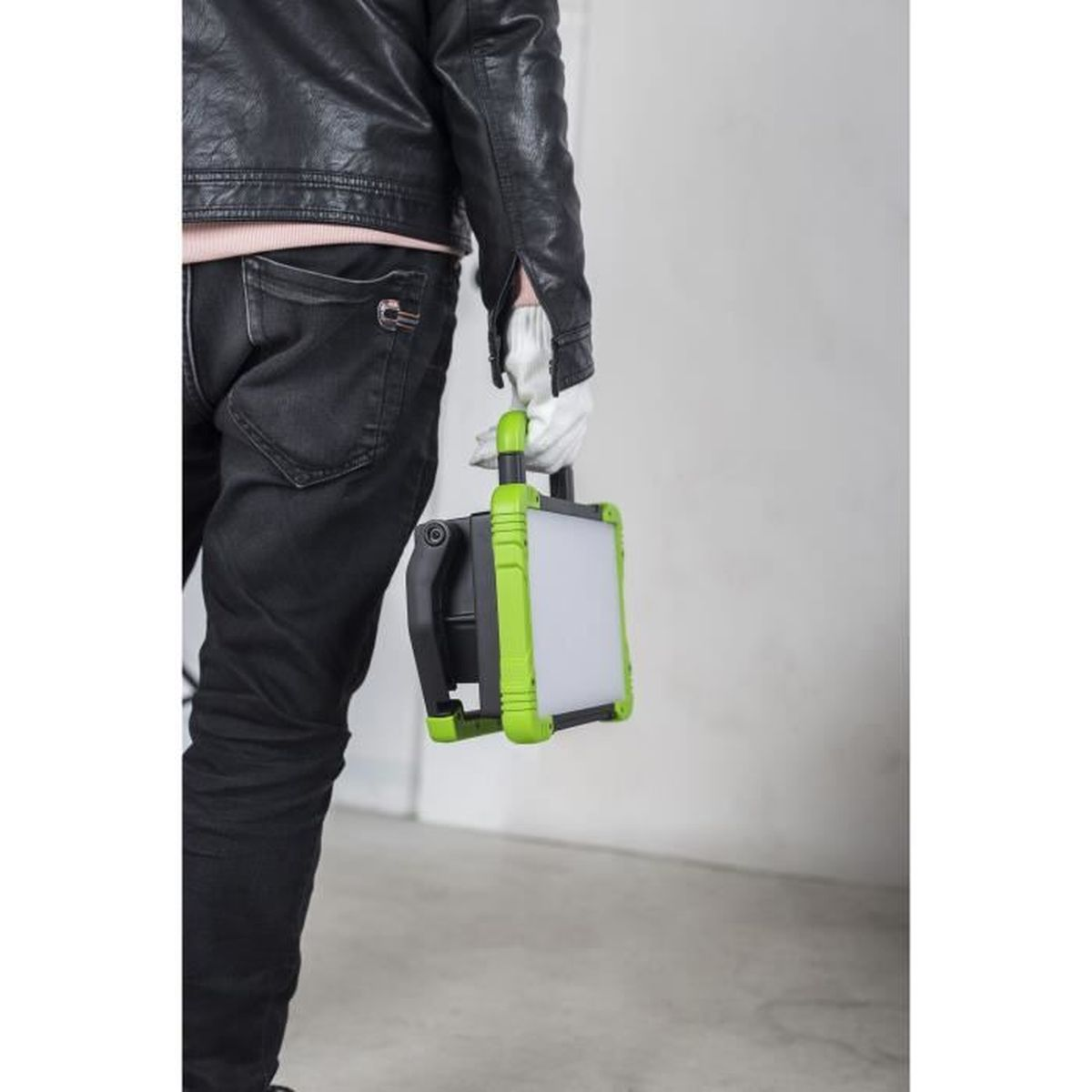 Person in schwarzer Jacke und Jeans trägt eine grüne und schwarze tragbare Arbeitsleuchte.