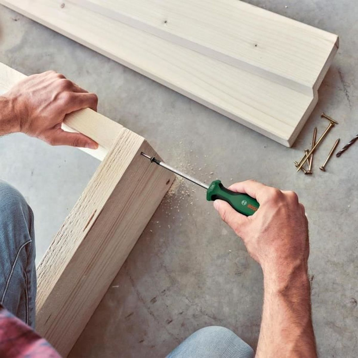 Eine Hand benutzt einen grünen Schraubendreher, um ein Holzstück auf ein anderes zu schrauben. Nägel und ein Bohrer liegen daneben.