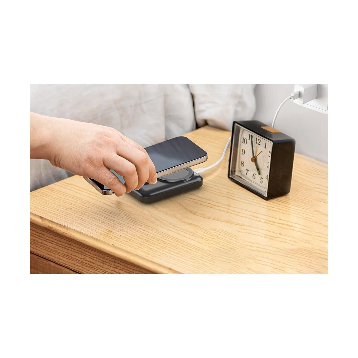 Eine Hand legt ein Smartphone auf ein Ladekabel neben einem Wecker.