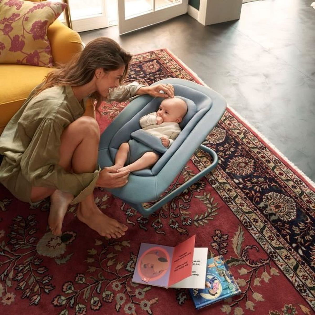 Eine Frau kniet neben einem Baby in einer blauen Wippe auf einem Teppich.