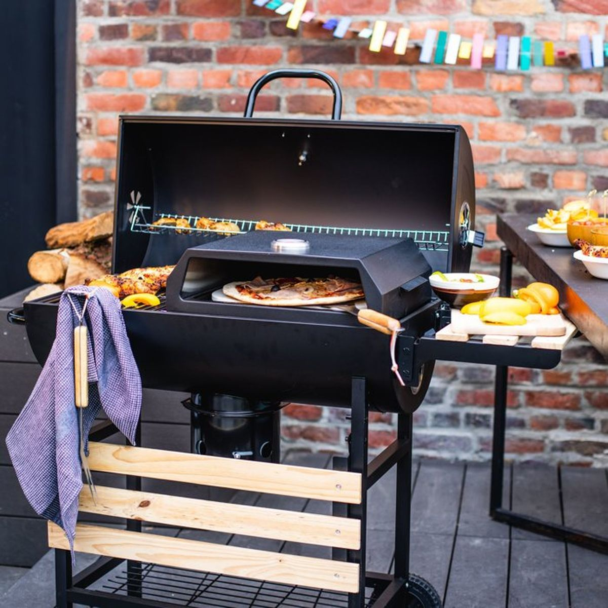 Ein schwarzer Grill mit einem Pizzastein darin, auf dem Pizza und Hähnchen garen. Holzscheite liegen daneben.