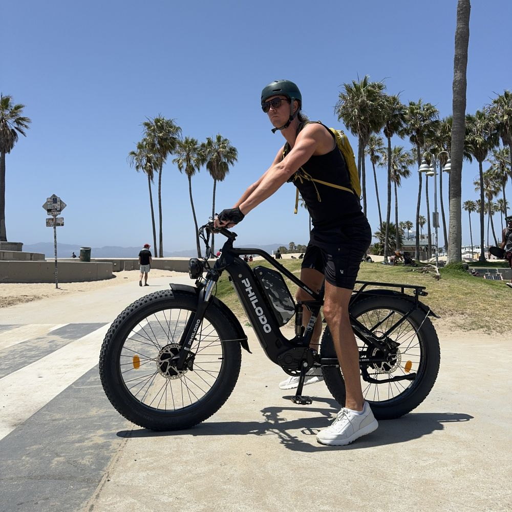 Uomo con casco su e-bike in spiaggia, palme sullo sfondo.