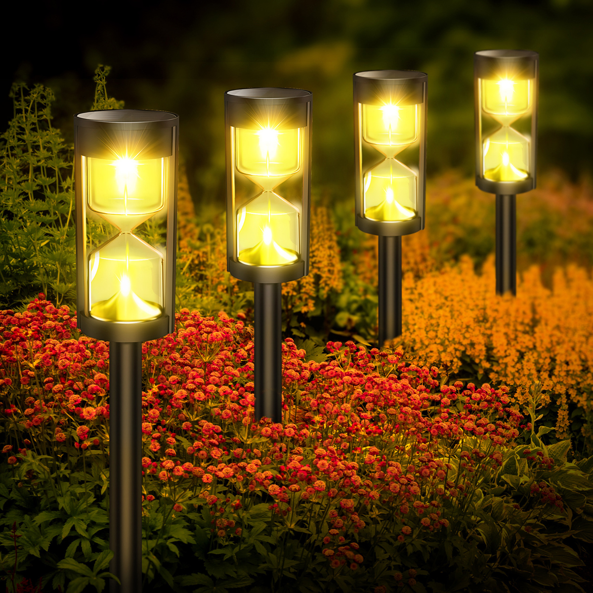 Vier sanduhrförmige Solarlichter leuchten gelb zwischen roten und orangen Blumen in der Dämmerung.