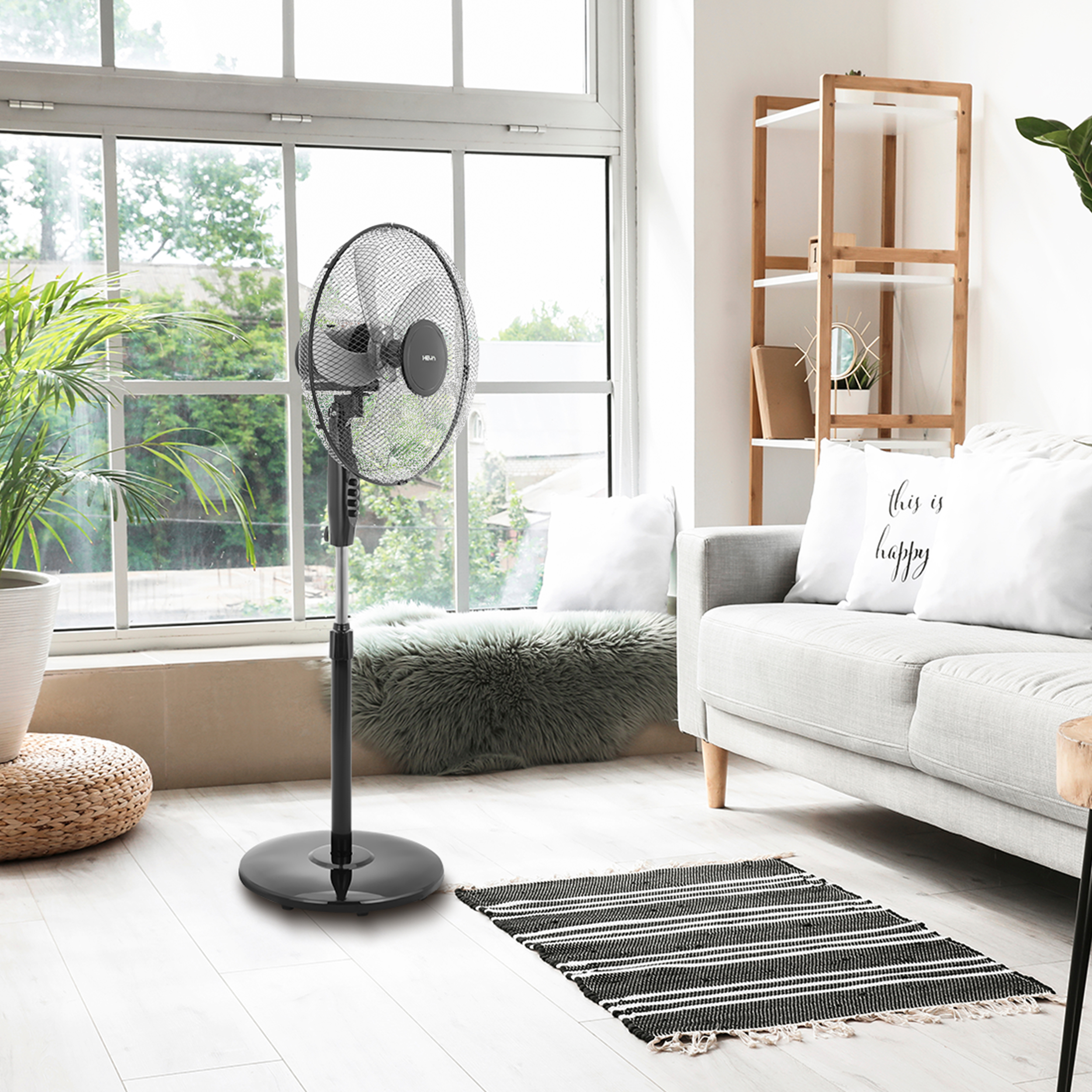 Czarny wentylator stojący znajduje się przed dużym oknem z widokiem na zieleń.