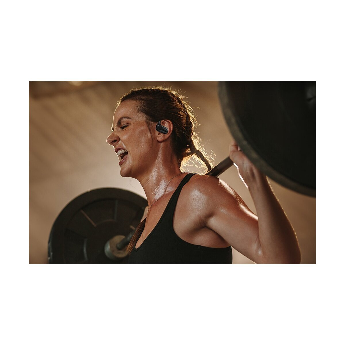 Eine verschwitzte Frau stemmt eine Hantel im Fitnessstudio, trägt Kopfhörer.