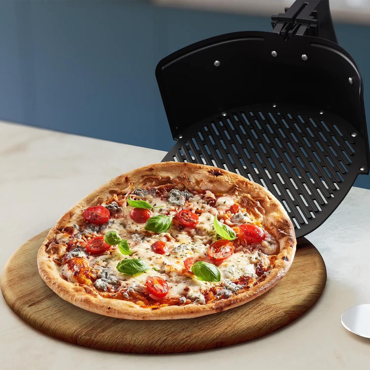 Eine heiße Pizza mit Tomaten und Basilikum auf einem Holzbrett neben einem Airfryer-Korb.