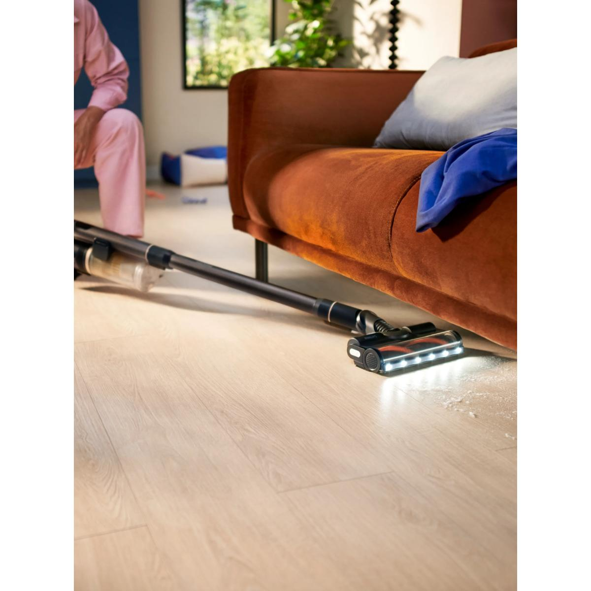Odkurzacz bezprzewodowy czyści drewnianą podłogę pod brązową kanapą.