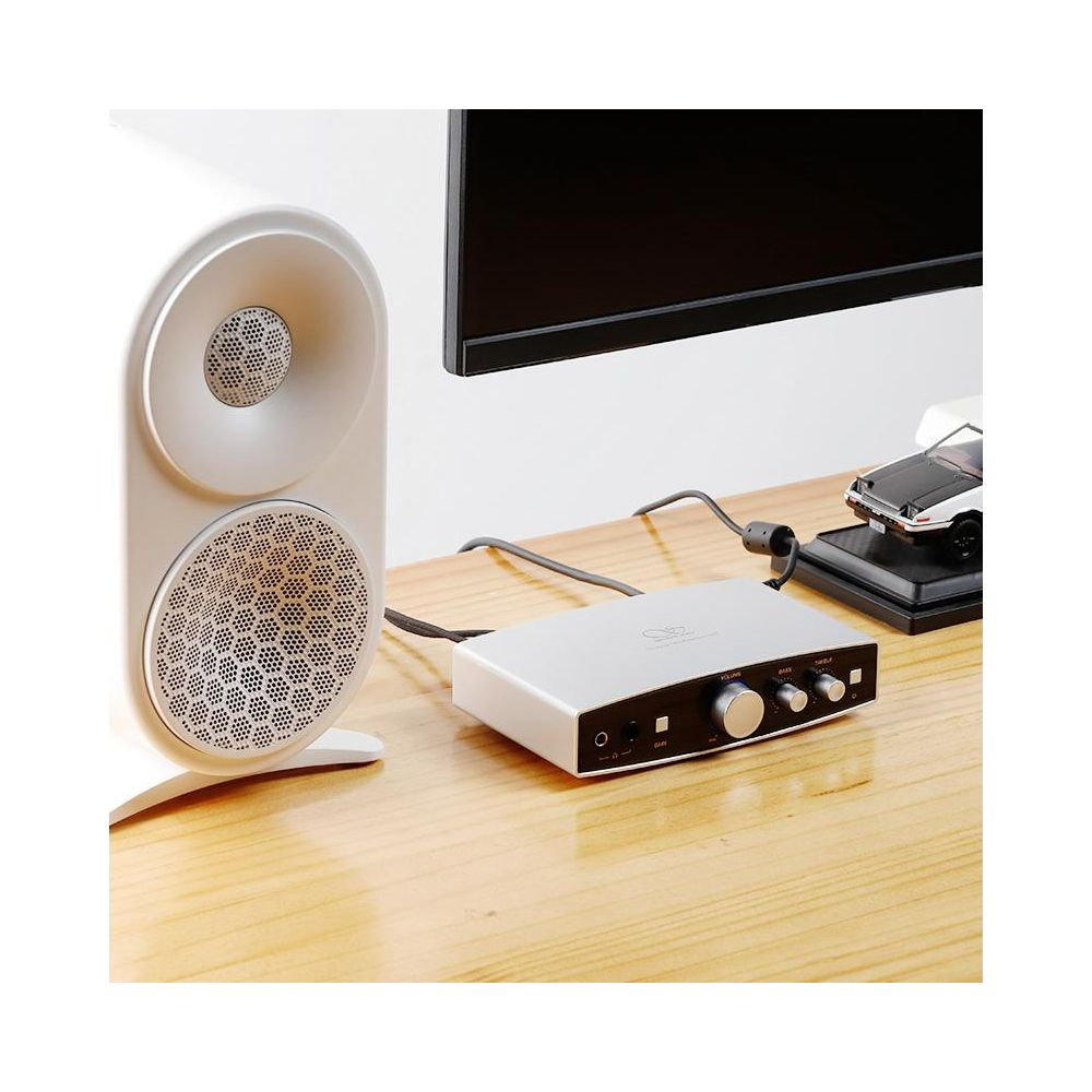 Un altavoz blanco, un dispositivo de audio plateado y un coche de juguete en un escritorio de madera.