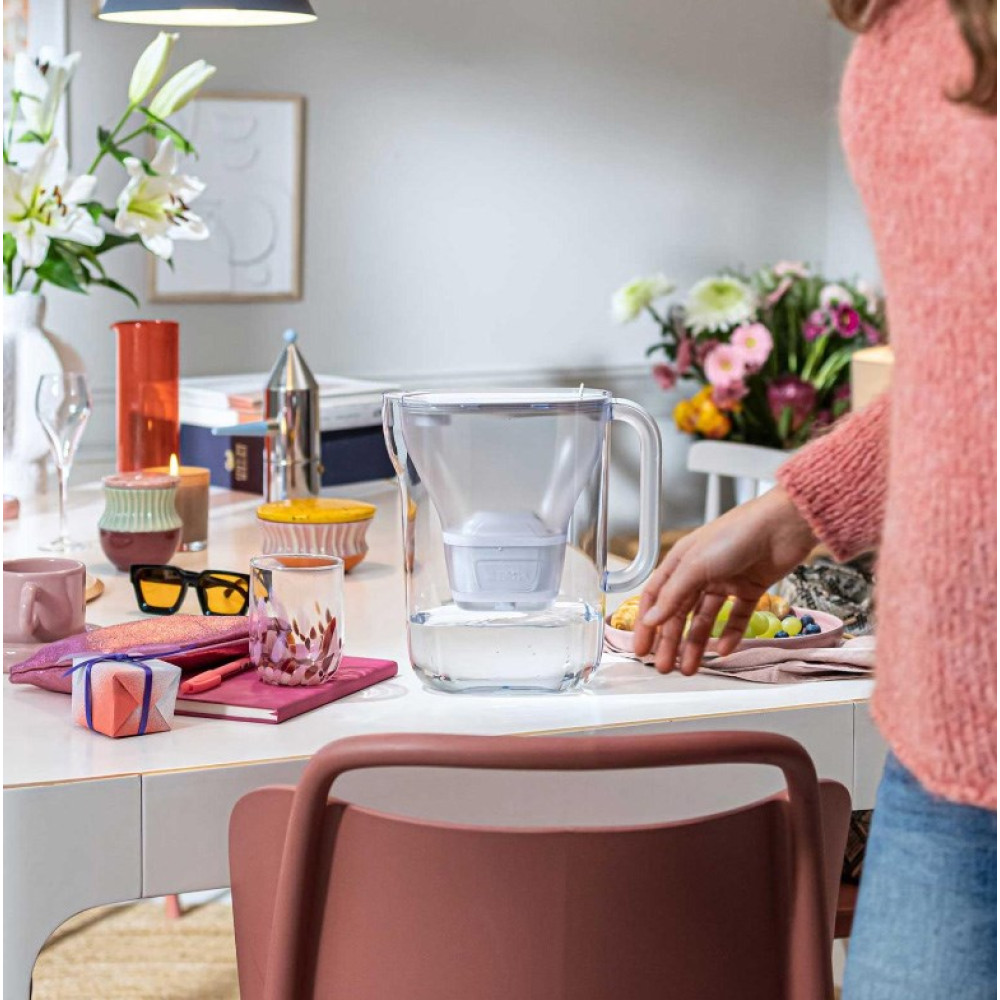 Osoba w różowym swetrze sięga po dzbanek filtrujący wodę na stole.