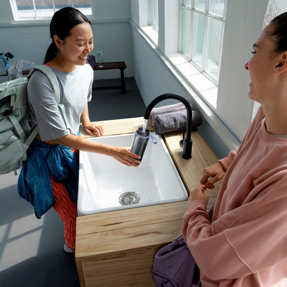 Dwie kobiety napełniają butelkę z wodą pod kranem w umywalce.