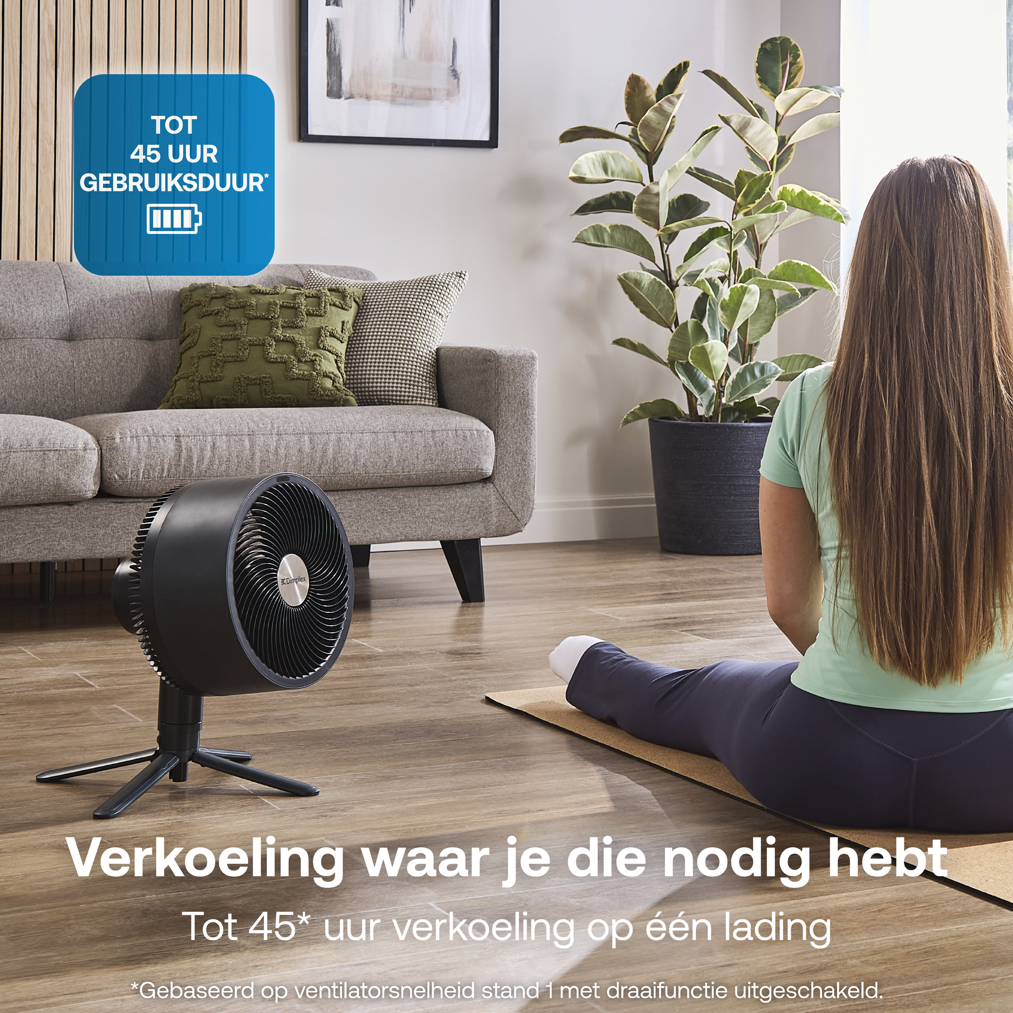 Een zwarte ventilator staat op een houten vloer naast een bank. Een vrouw zit op een mat.