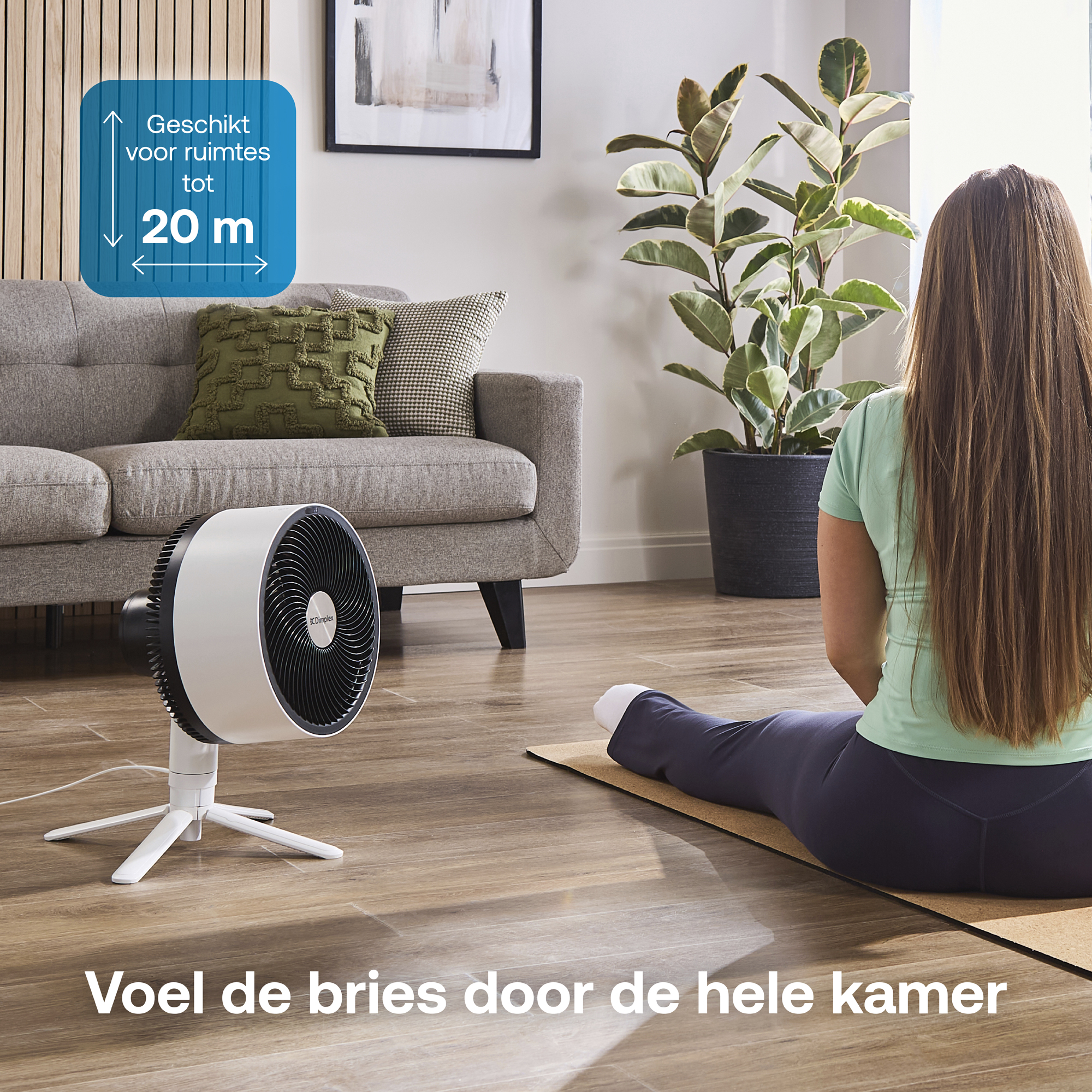 Een ventilator staat op de vloer voor een bank. Een vrouw zit met haar rug naar de camera op een mat.