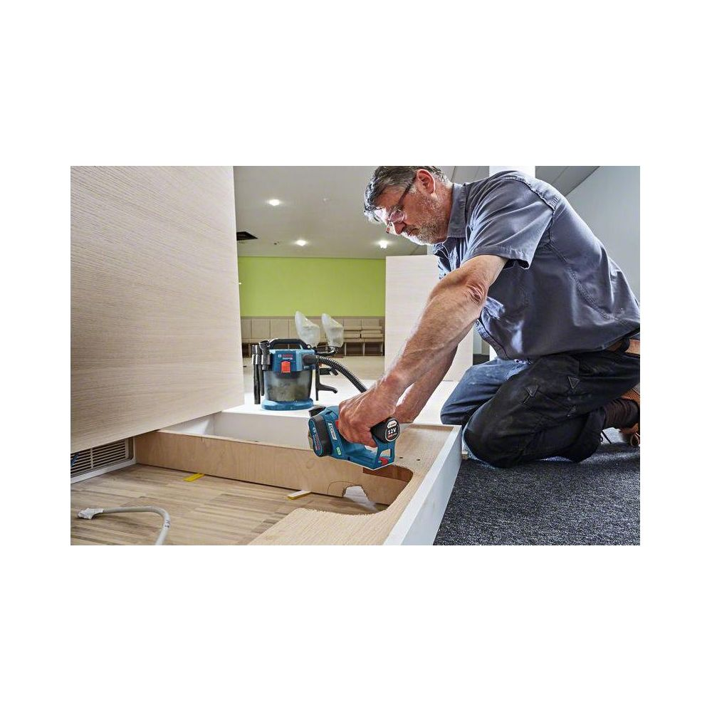 Mężczyzna klęczy, używając frezarki do drewna. Obok stoi odkurzacz.