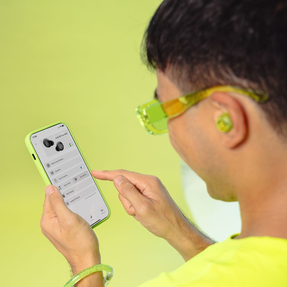 Mann im gelben Hemd benutzt Handy mit Ohrhörer-App, trägt grüne Brille und Ohrhörer.