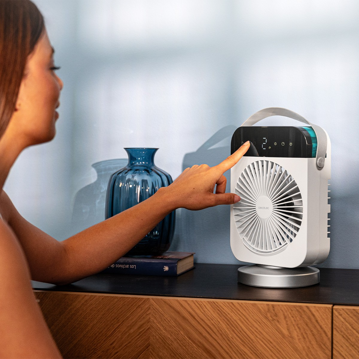 Mujer operando un ventilador blanco con pantalla, junto a un jarrón azul.