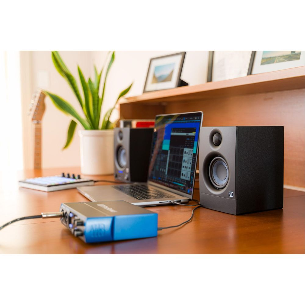 Un laptop, interfaccia audio e altoparlanti su scrivania in legno con pianta e chitarra.