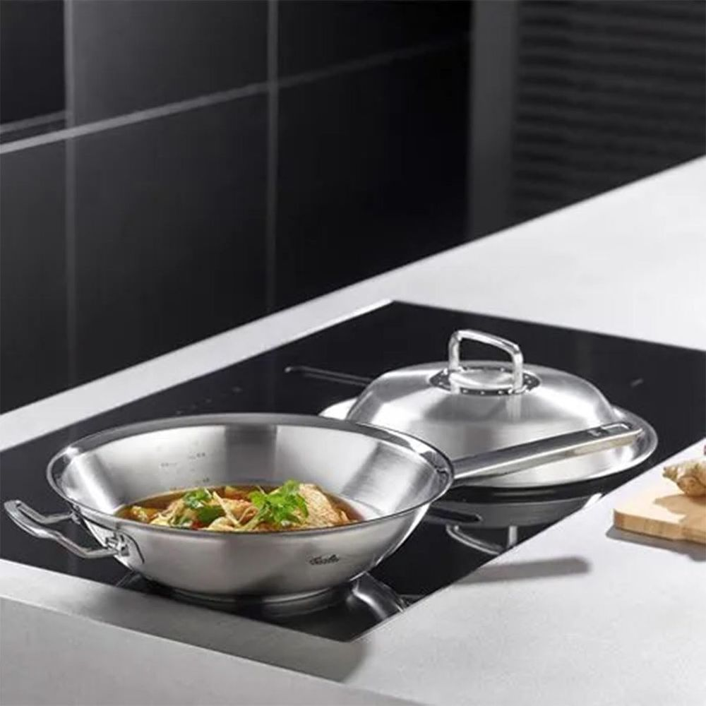 Un wok in acciaio inossidabile con cibo si trova su un piano cottura accanto a una pentola con coperchio.