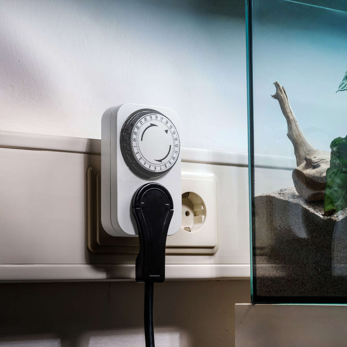 Een witte timerstekker met zwart snoer naast een stopcontact en een aquarium.