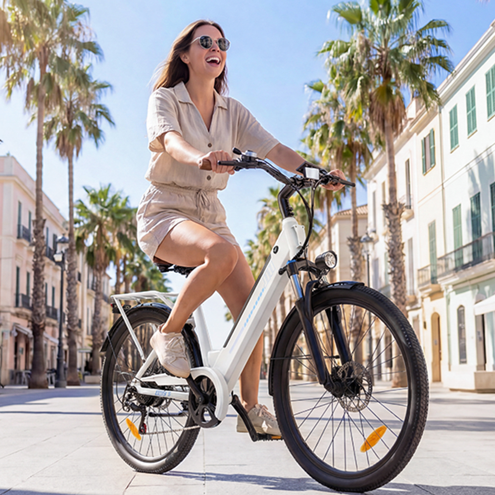 Frau fährt weißes E-Bike auf palmengesäumter Straße mit Gebäuden im Hintergrund.
