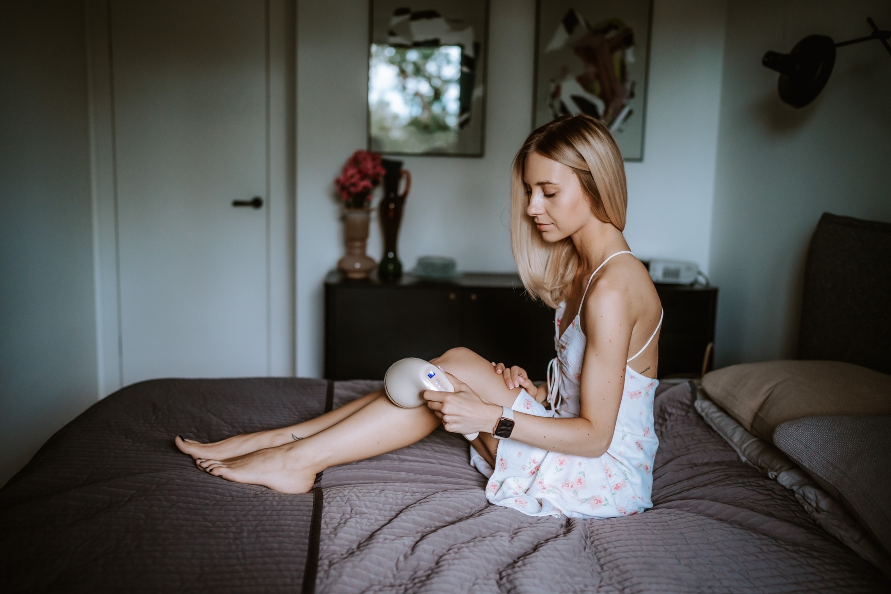 Eine blonde Frau in einem geblümten Kleid benutzt ein weißes Haarentfernungsgerät an ihrem Bein, während sie auf einem Bett sitzt.