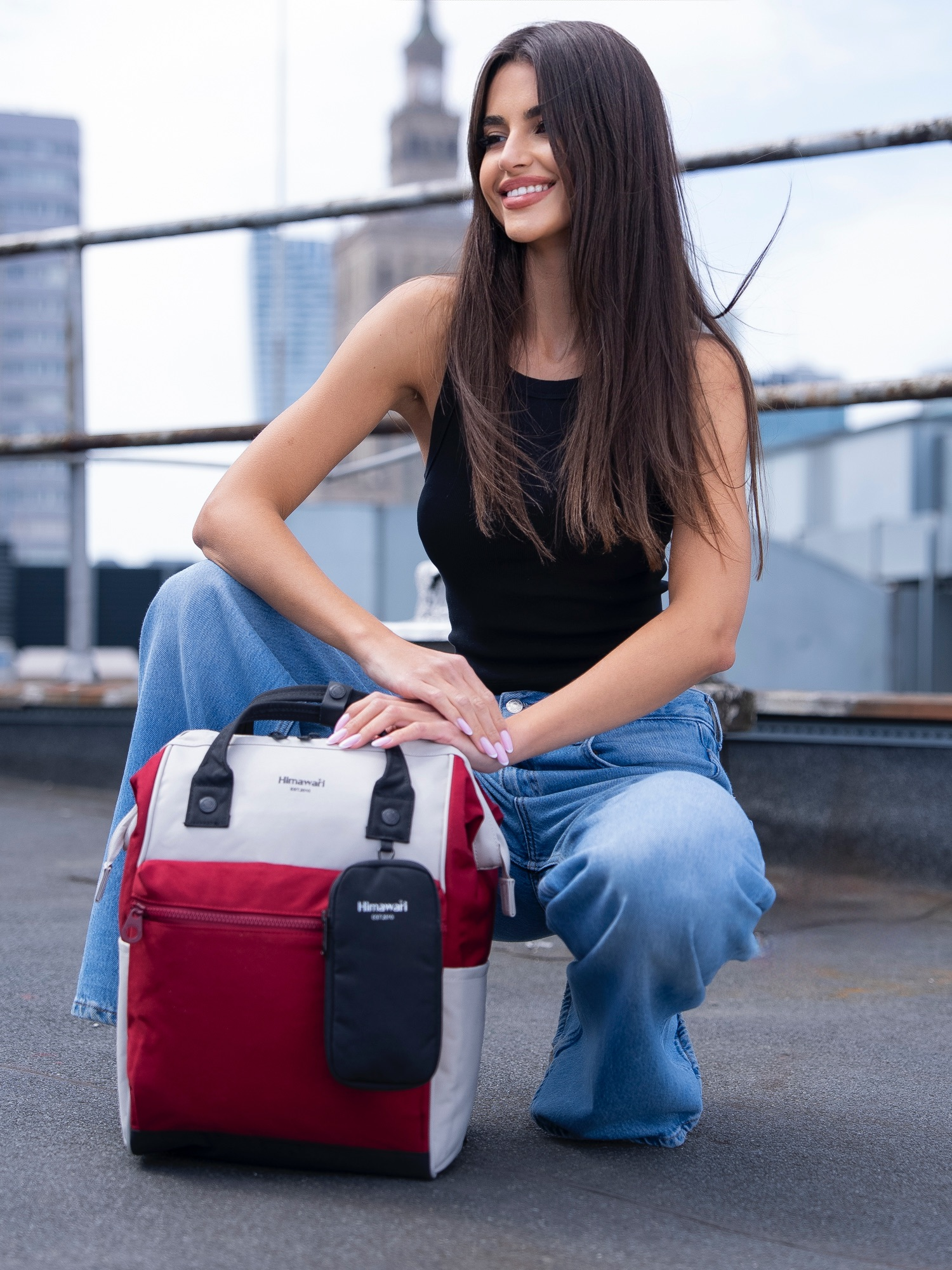 Kobieta w dżinsach i czarnym topie klęczy obok czerwono-białego plecaka na dachu.
