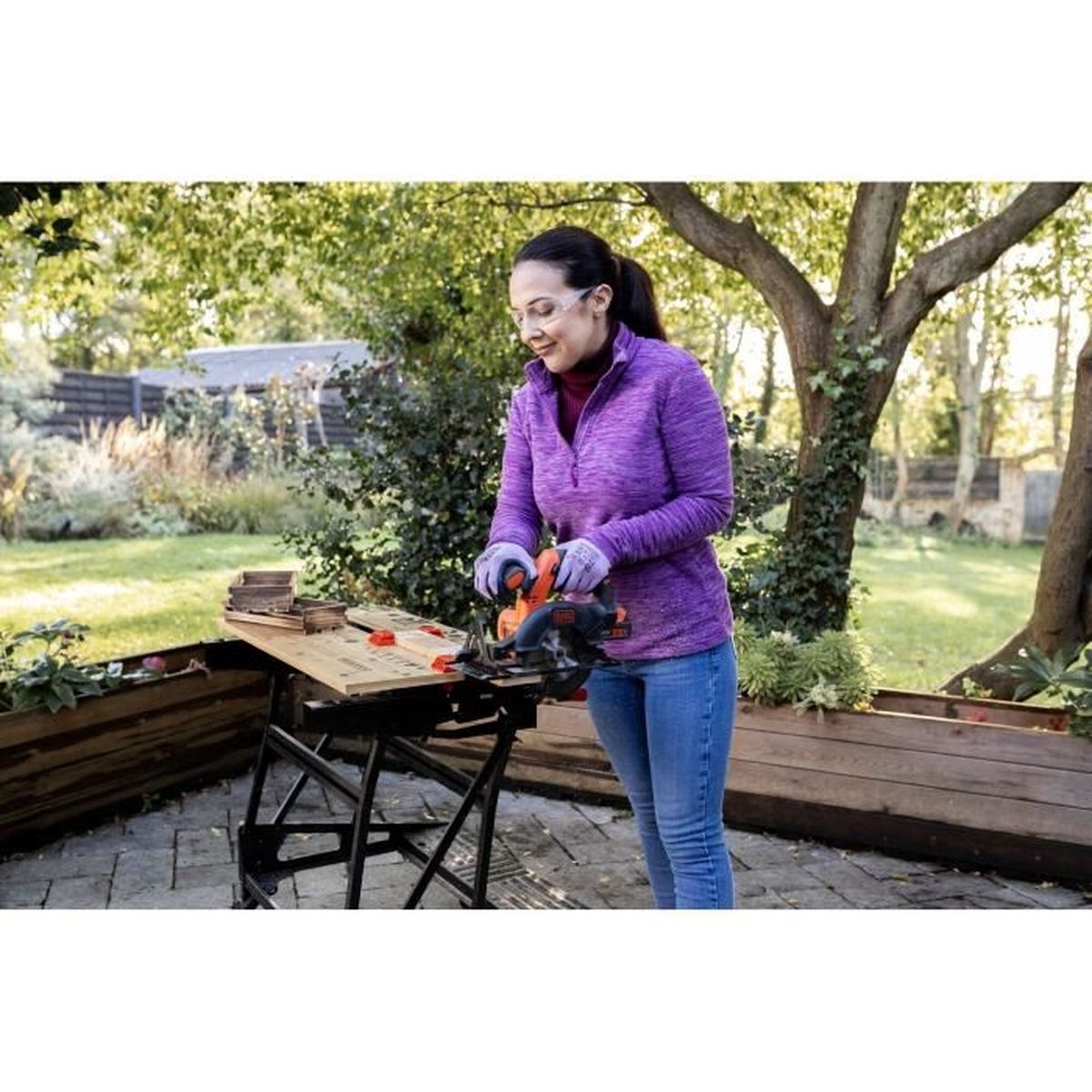 Kobieta w okularach ochronnych tnie drewno na koziołku w ogrodzie.