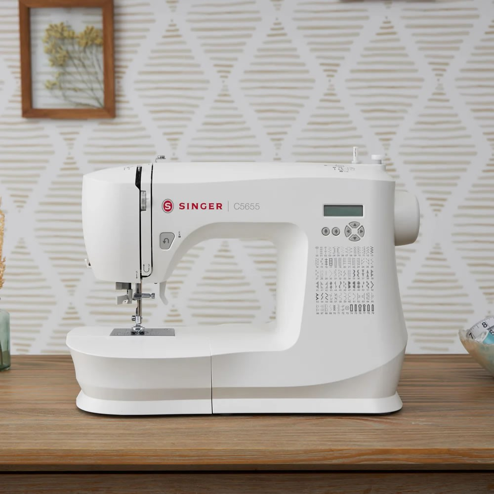Beyaz Singer C5655 dikiş makinesi, desenli duvar kağıdı önündeki ahşap masada duruyor.