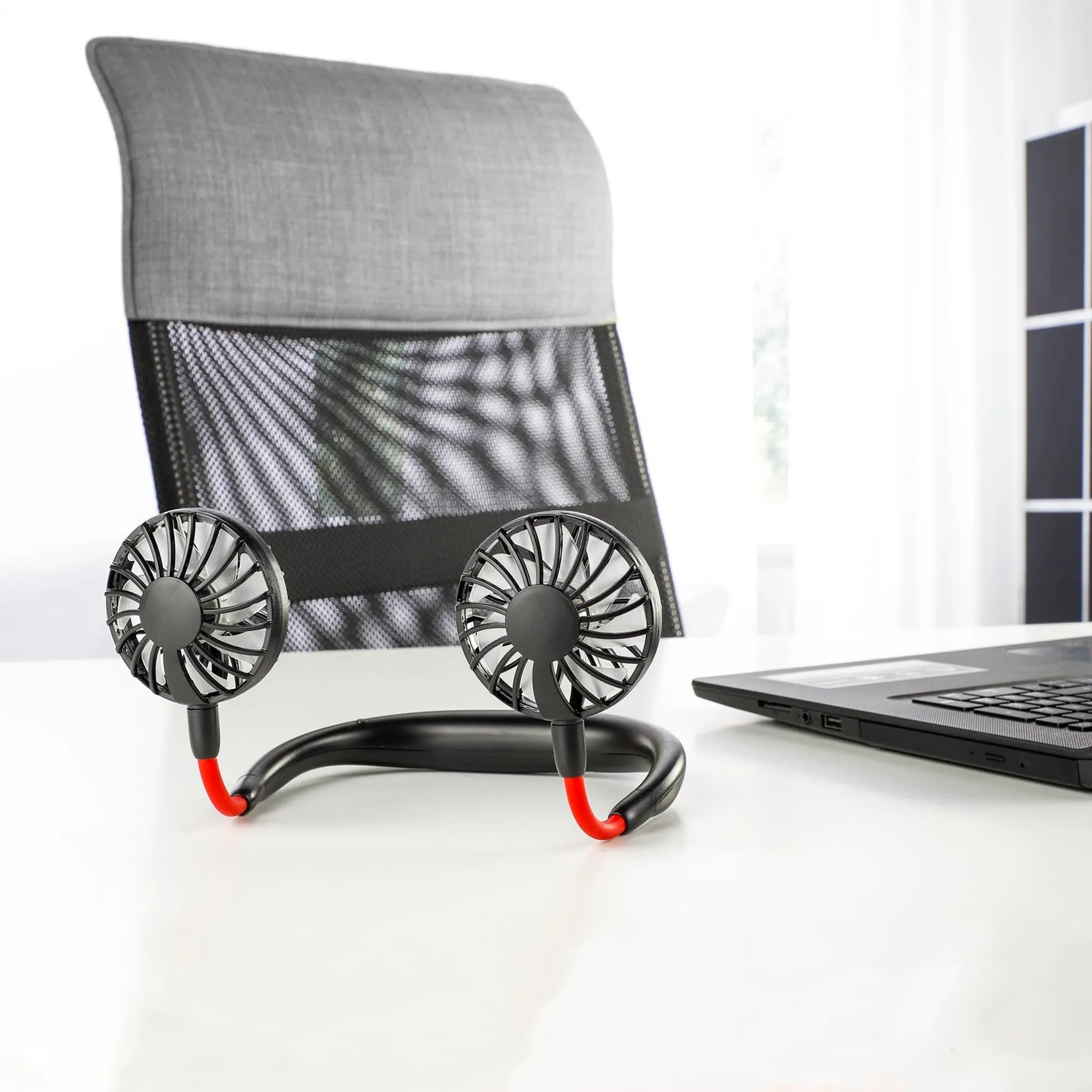 Ein Nackenventilator mit zwei schwarzen Lüftern steht auf einem weißen Schreibtisch neben einem Laptop.