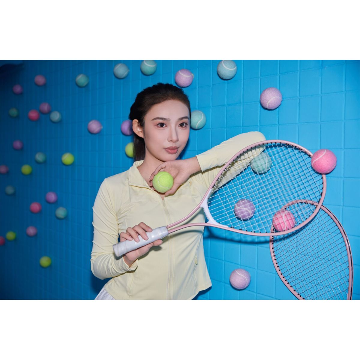 Woman holding tennis rackets and colorful balls against a blue wall.