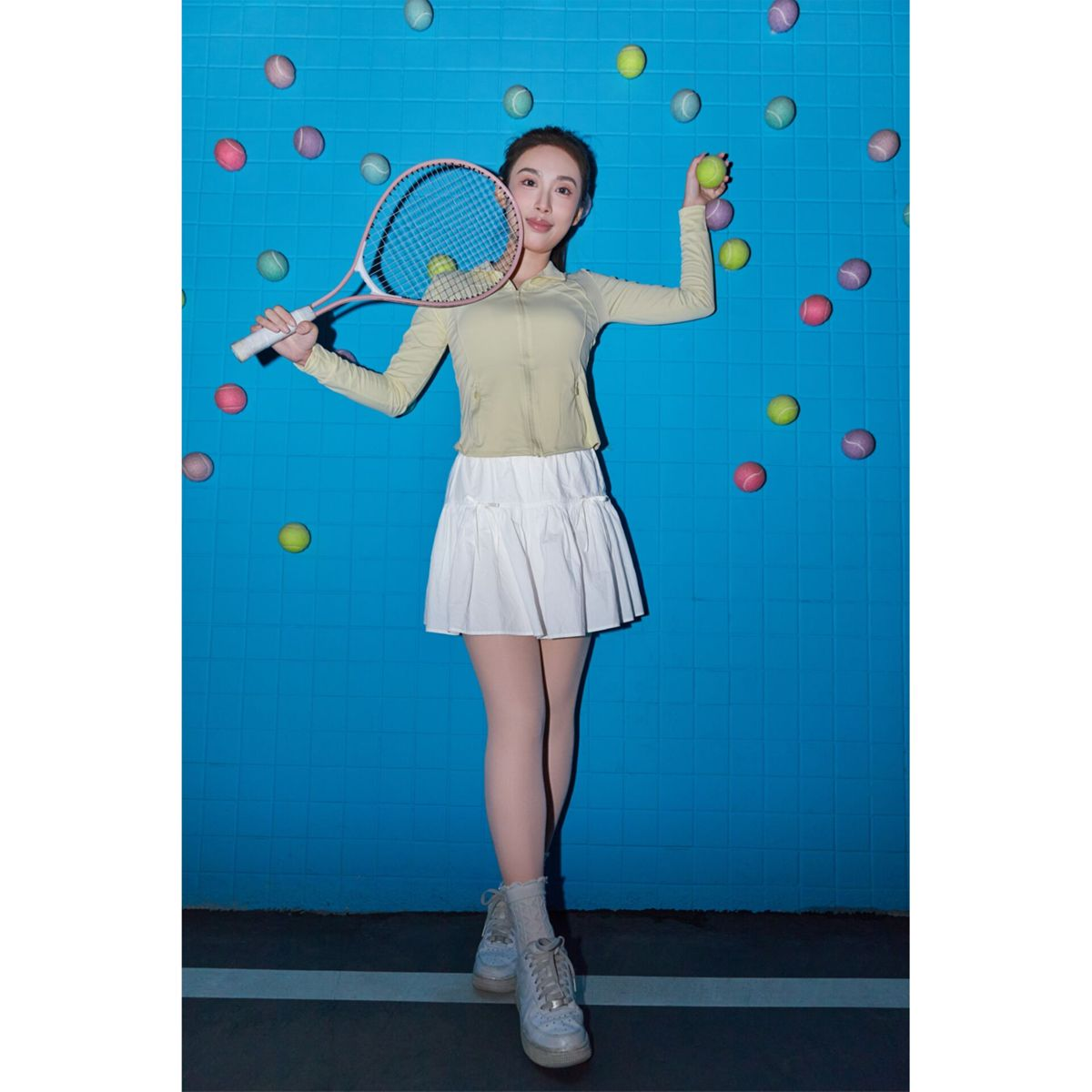 A woman in tennis attire holds a racket and ball against a blue background with tennis balls.