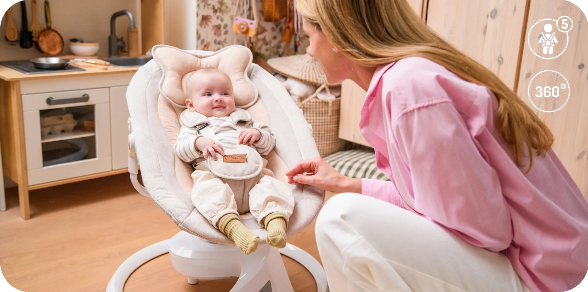 Eine Frau in einem rosa Hemd schaut auf ein Baby in einer Wiege mit Spielzeugküche im Hintergrund.