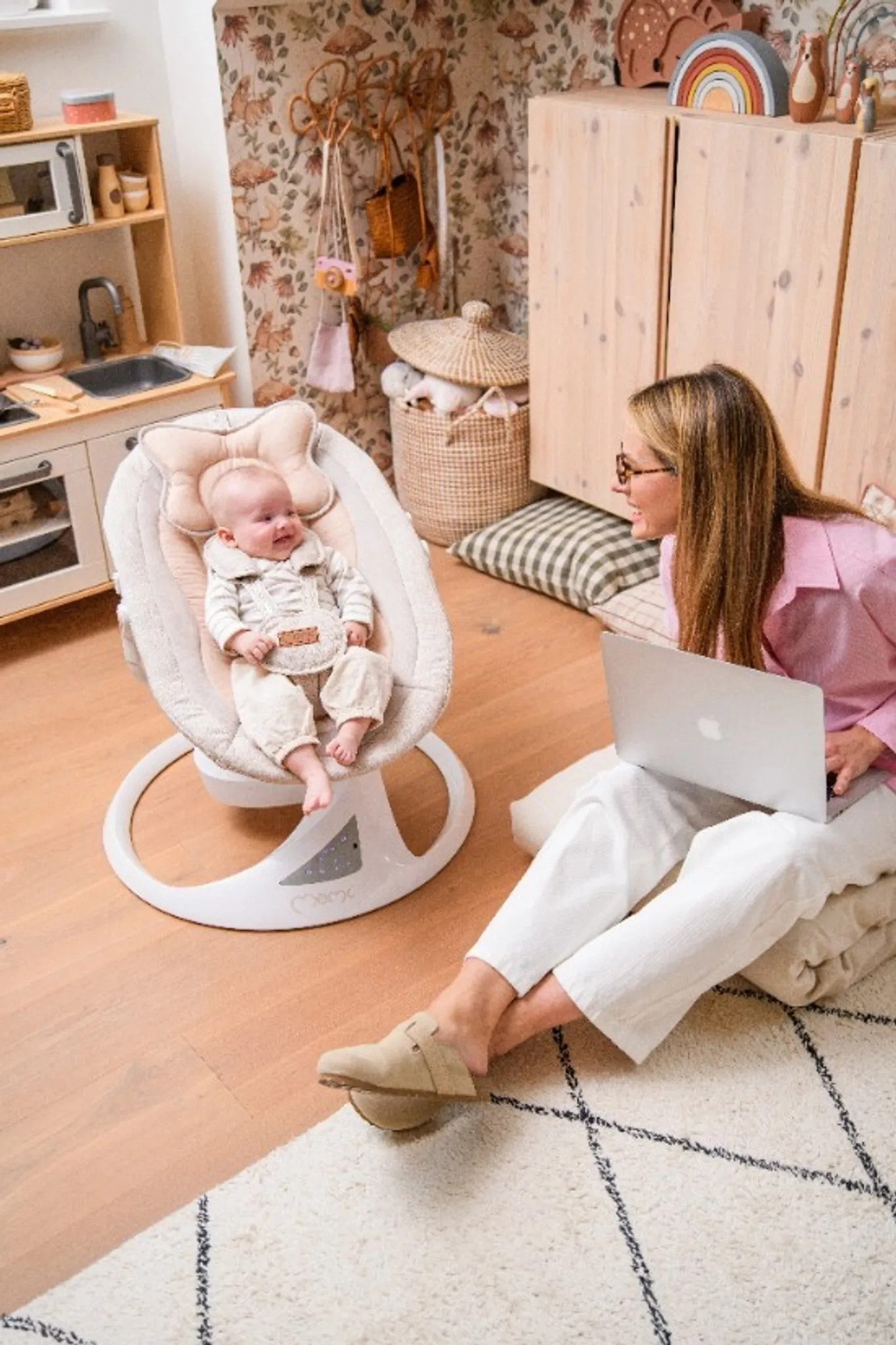 Eine Frau mit Laptop sitzt auf dem Boden vor einem Baby in einer Babywippe.