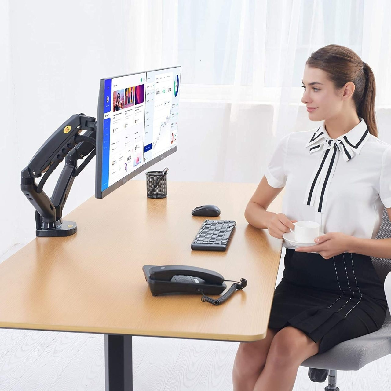 Kadın beyaz gömlek ve siyah etekle, çift monitörlü, telefonlu, klavyeli ve fareli masada.